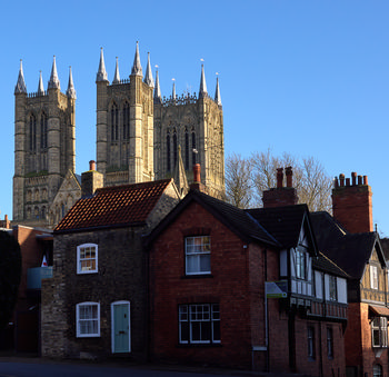In this urban architectural photograph, Lincoln Cathedral rises prominently above a row of traditional houses in Lincoln, England. The iconic towers and spires of Lincoln Cathedral, a famous landmark in the United Kingdom, are illuminated by the clear midday winter sunlight, casting sharp lines against the blue sky. The setting, with its mix of historic red brick and stone houses in the foreground, highlights the rich architectural heritage of Lincoln, while the cathedral dominates the skyline as a testament to the city’s medieval past. The image captures the crisp, bright atmosphere characteristic of a winter afternoon in England, making Lincoln Cathedral the unmistakable focal point of this picturesque scene.
